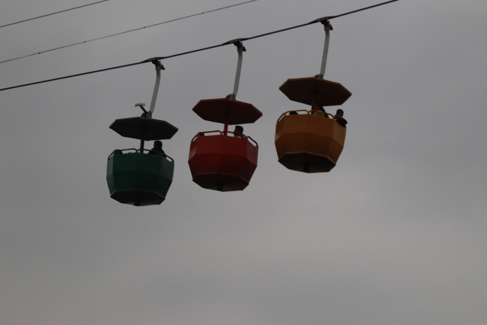 Three cable cars suspended on overhead cables, each carrying passengers, are shown against a cloudy sky.