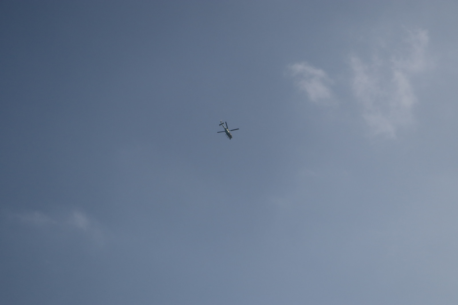 A helicopter flies against a clear blue sky with a few scattered clouds.